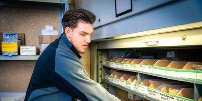 Ein junger Mann sortiert sorgfältig Kartons in einem Regal, umgeben von verschiedenen beschrifteten Boxen und Materialien.