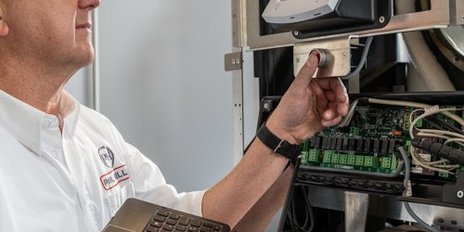 Ein Techniker mit Brille hält ein Laptop und überprüft ein Gerät in einem technischen Umfeld mit vielen Kabeln.