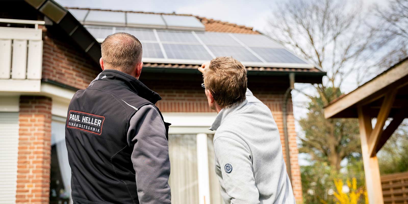 Zwei Männer betrachten die Solarpanels auf einem Dach, während sie sich angeregt unterhalten und auf die Technik zeigen.