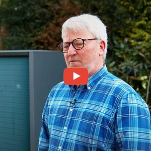 Ein älterer Mann mit Brille trägt ein kariertes Hemd und schaut nachdenklich in die Kamera, während er im Freien steht.