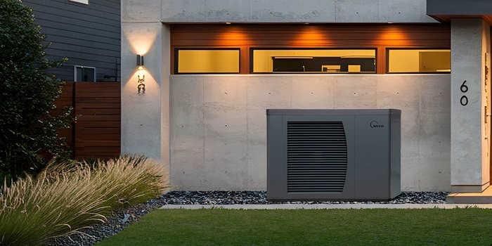 Ein modernes Haus mit einer grauen Wärmepumpe, die vor einer Betonwand und großen Fenstern platziert ist.