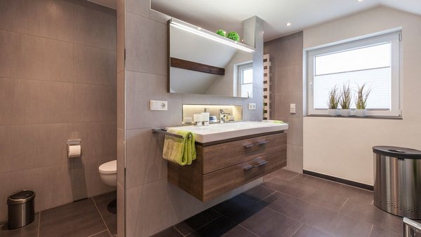 Ein modernes Badezimmer mit einem schwebenden Waschbecken, Holzschubladen und einem großen Spiegel, der das Licht reflektiert.