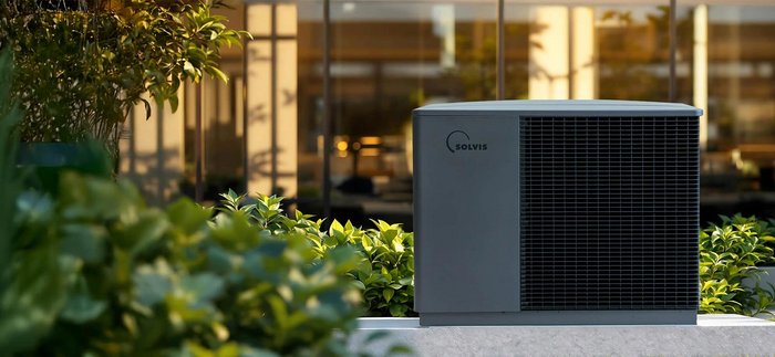 Eine moderne Klimaanlage steht in einem gepflegten Garten, umgeben von üppigem Grün und einem eleganten Gebäude im Hintergrund.