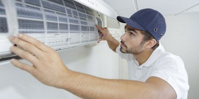 Ein Mechatroniker installiert eine Klimaanlage an der Wand und trägt eine Schirmmütze auf dem Kopf.