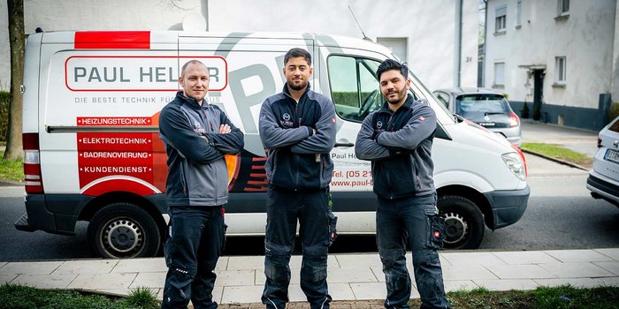 Drei Handwerker stehen selbstbewusst vor einem Firmenwagen, der mit Dienstleistungen wie Heizungs- und Elektrotechnik beworben wird.