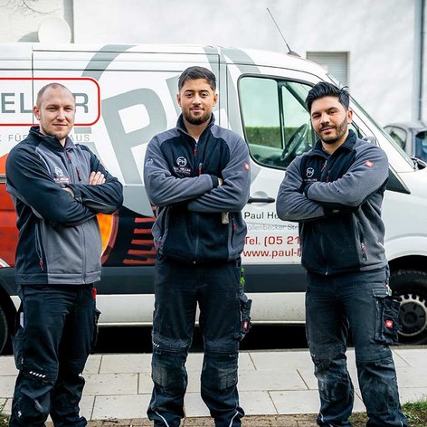 Drei Handwerker stehen selbstbewusst vor einem Firmenwagen, der mit Dienstleistungen wie Heizungs- und Elektrotechnik beworben wird.