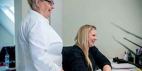 Eine Frau mit langen Haaren tippt an einem Computer, während eine andere Frau in einem weißen Hemd interessiert zuschaut.
