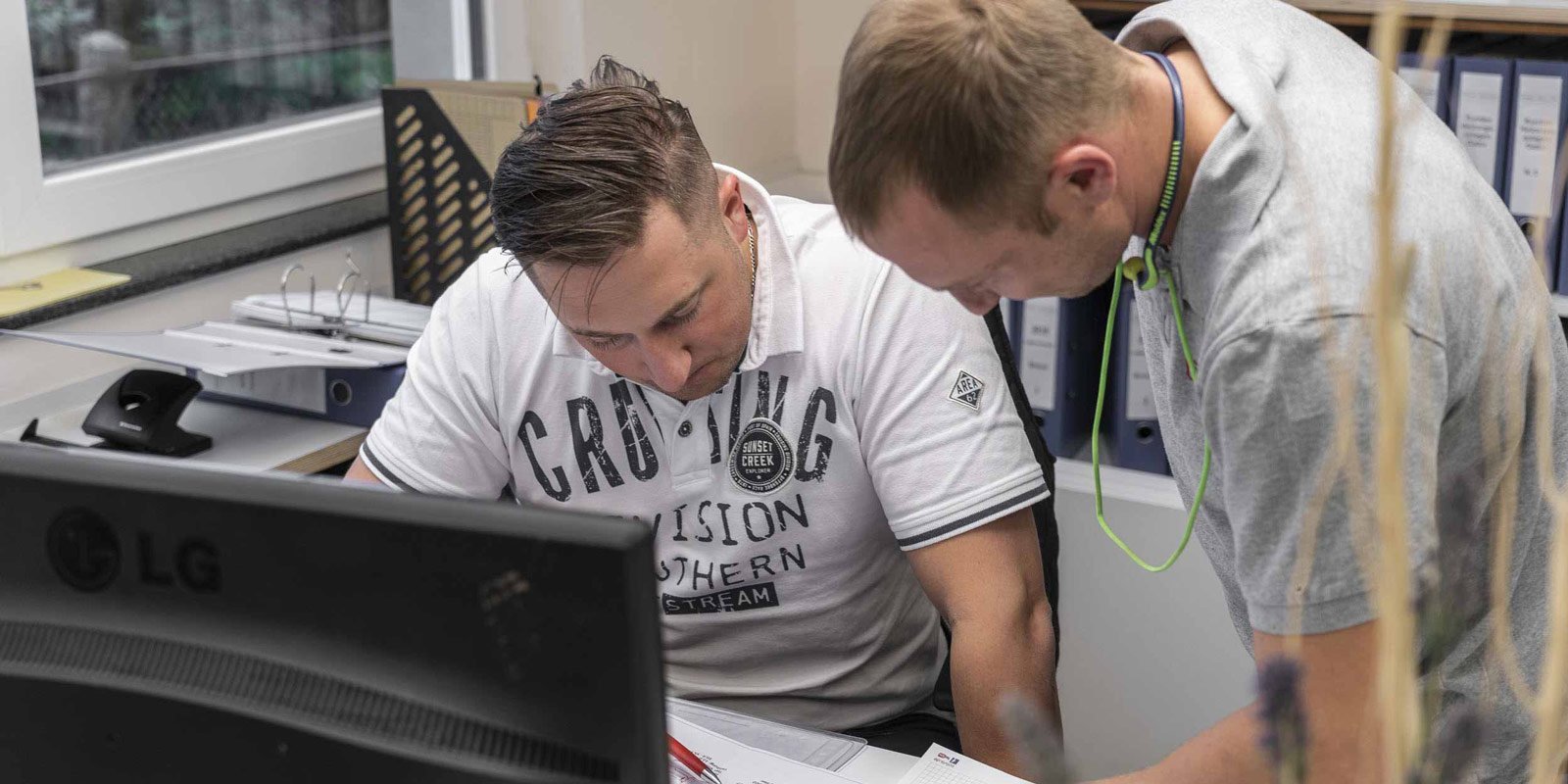 Zwei Männer konzentrieren sich auf Dokumente am Schreibtisch, während ein Computer im Vordergrund sichtbar ist.