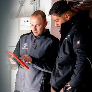 Zwei Männer betrachten gemeinsam ein Tablet, während sie in einem technischen Raum stehen, umgeben von Kabeln und Geräten.