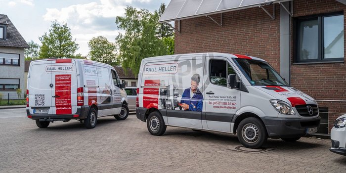 Zwei Lieferwagen der Firma Paul Heller stehen auf einem gepflasterten Parkplatz vor einem modernen Gebäude.
