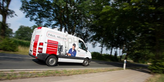 Ein Lieferwagen mit der Aufschrift 'Paul Heller' fährt auf einer Landstraße, umgeben von Bäumen und grünem Gras.