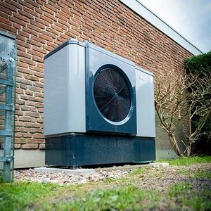Eine moderne Wärmepumpe steht an einer Backsteinwand, umgeben von einem gepflegten Garten mit kleinen Steinen.
