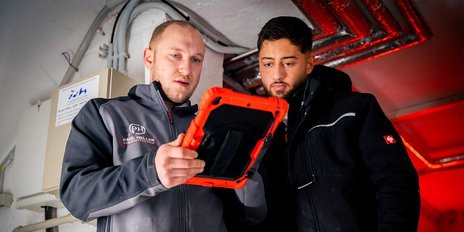 Zwei Männer betrachten konzentriert ein Tablet, während sie in einem technischen Raum mit rotem Licht stehen.