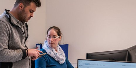 Ein Mann zeigt einer Frau auf seinem Smartphone etwas, während sie an einem Schreibtisch sitzt und aufmerksam schaut.