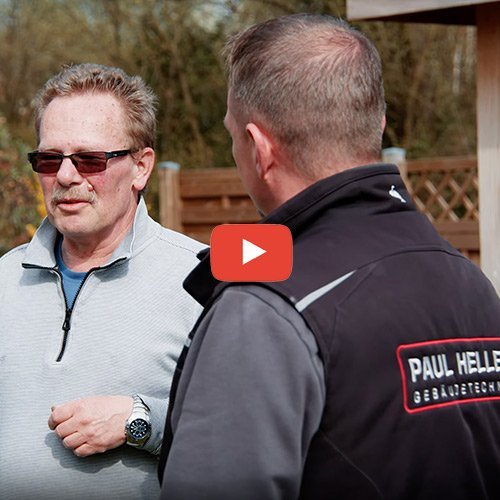 Zwei Männer unterhalten sich im Freien, einer trägt eine Sonnenbrille und einen grauen Pullover, der andere eine schwarze Jacke.