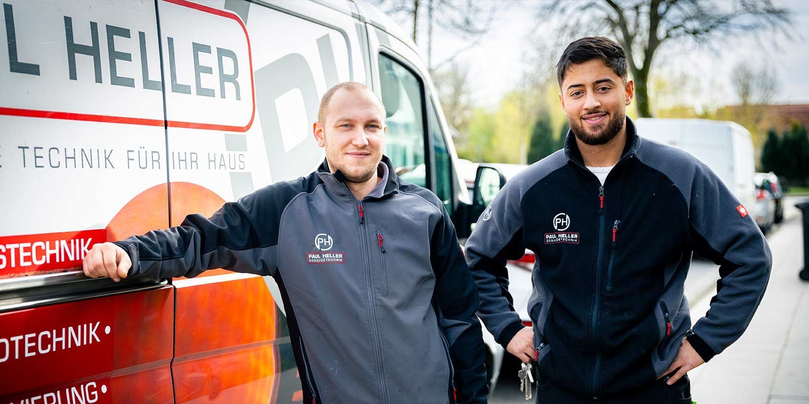 Zwei Männer in Arbeitskleidung stehen lächelnd vor einem Fahrzeug mit dem Logo von Paul Heller, umgeben von einer grünen Umgebung.
