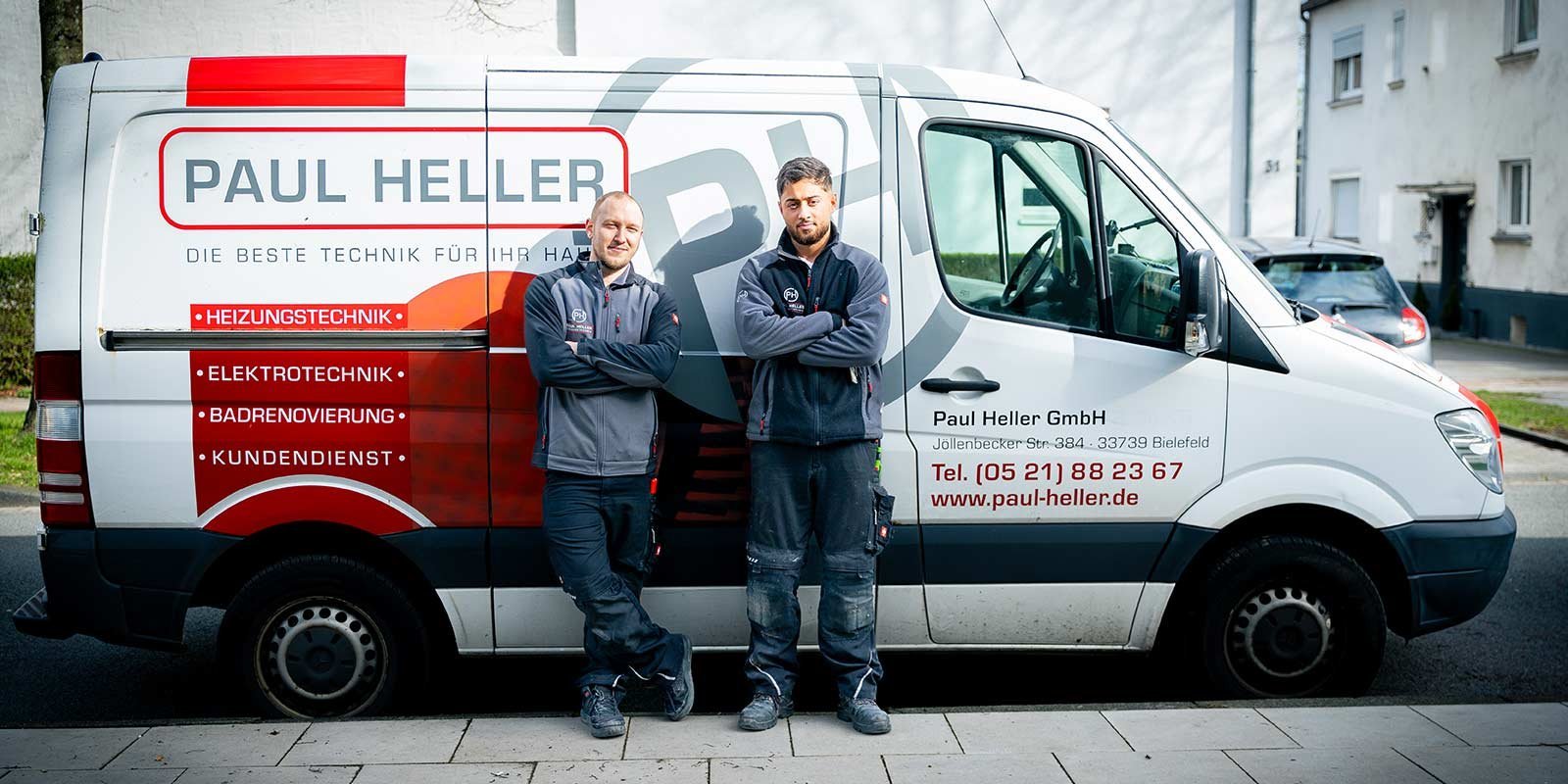 Zwei Techniker stehen lässig vor einem Firmenwagen, der mit dem Logo und den Dienstleistungen von Paul Heller beschriftet ist.