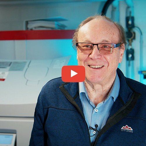 Der Mann trägt eine blaue Jacke und hat eine entspannte Ausstrahlung, umgeben von technischen Geräten im Hintergrund.