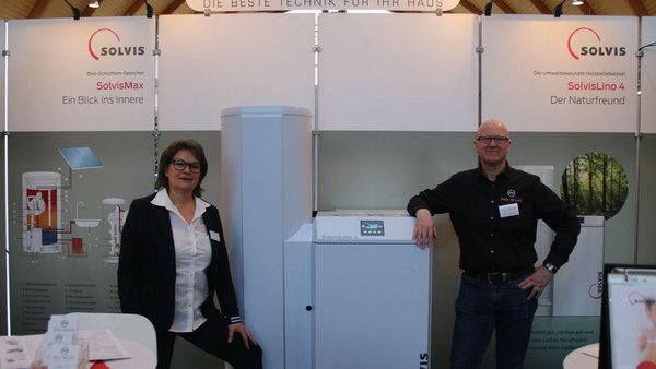 Eine Frau in einem weißen Hemd und ein Mann in schwarzem Hemd präsentieren Heizsysteme auf einer Messe, lächelnd und freundlich.