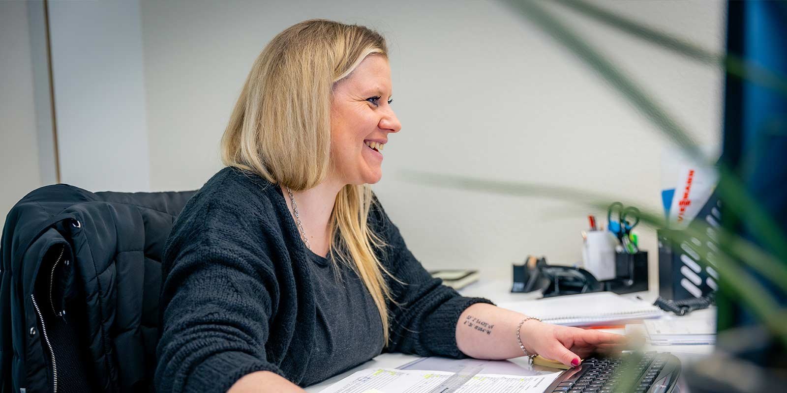 Eine lächelnde Frau mit langen blonden Haaren arbeitet an ihrem Schreibtisch und tippt auf einer Tastatur.