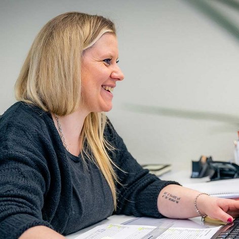 Eine lächelnde Frau mit langen blonden Haaren arbeitet an ihrem Schreibtisch und tippt auf einer Tastatur.