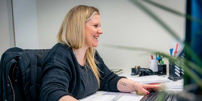 Eine lächelnde Frau mit langen blonden Haaren arbeitet an ihrem Schreibtisch und tippt auf einer Tastatur.
