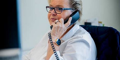 Eine Frau mit blonden, kurzen Haaren und Brille spricht am Telefon, während sie an einem Schreibtisch sitzt.