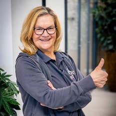 Eine lächelnde Frau mit Brille zeigt ein Daumen-hoch-Zeichen in einem modernen Innenraum mit Pflanzen im Hintergrund.